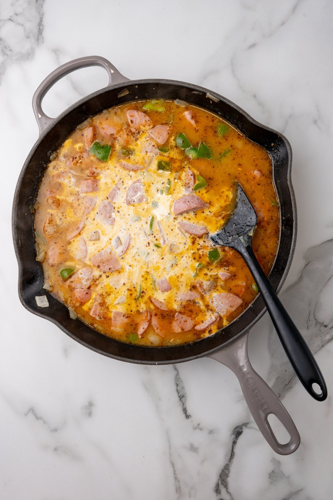 Top down view of a skillet with cream added into kielbasa, green pepeprs and onions