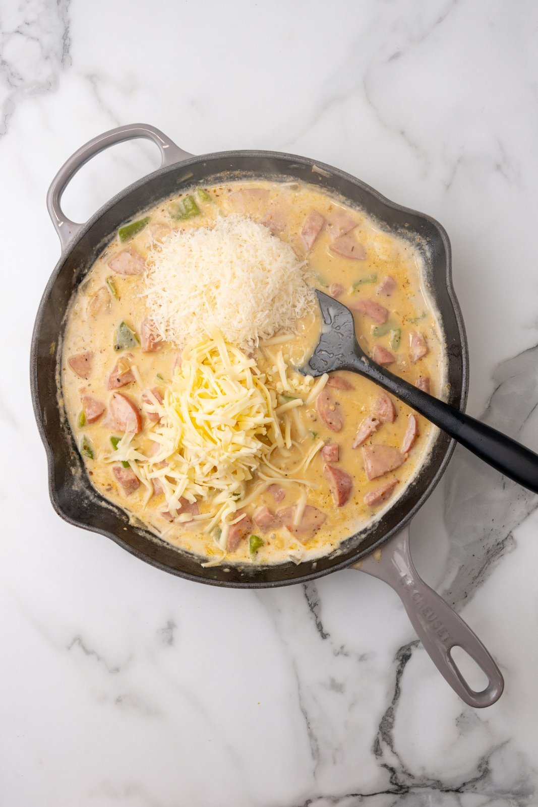 Top down view of shredded cheddar and parmesean cheese added to a skillet cooking kielbasa, green pepeprs and onions