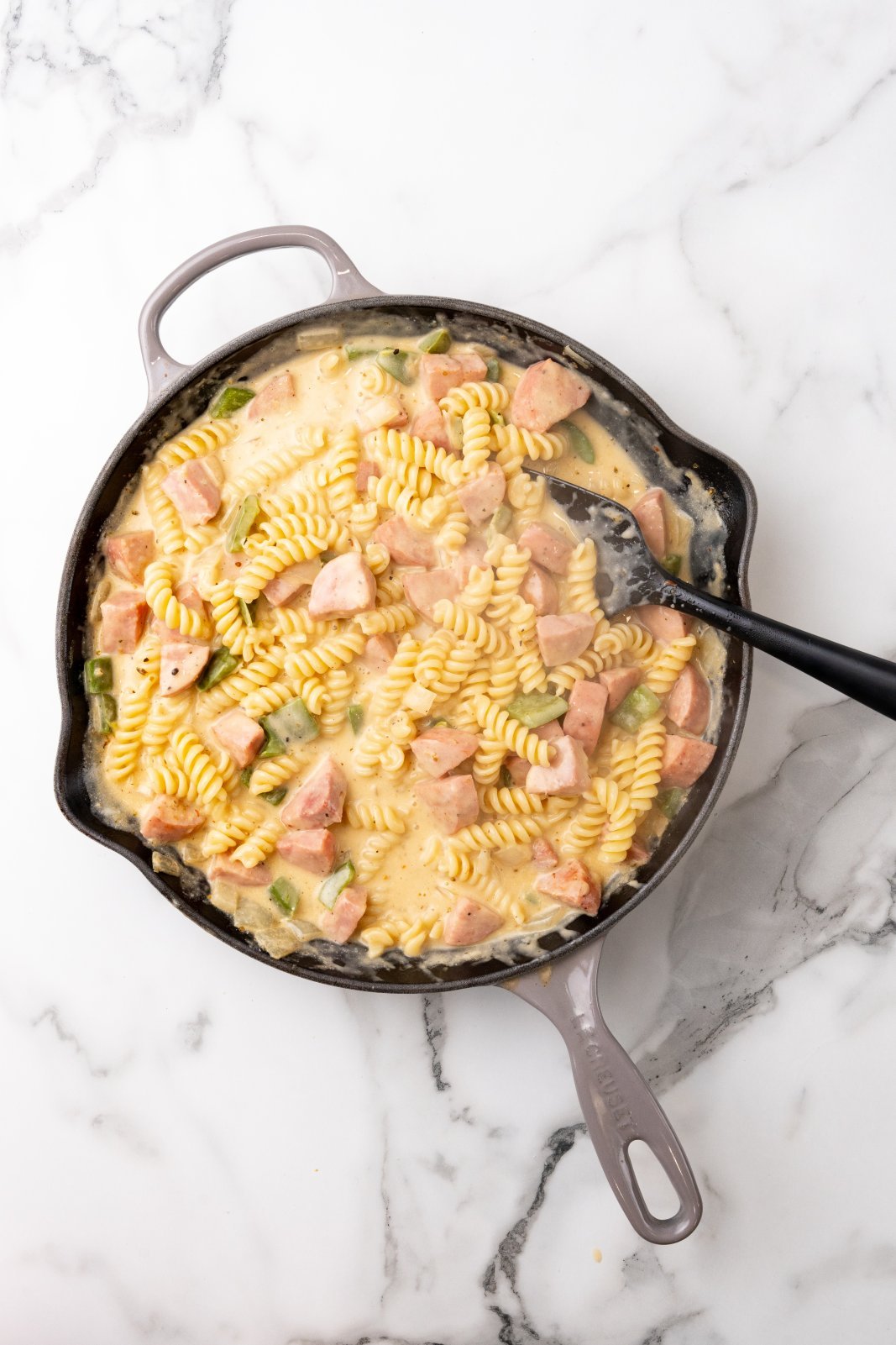 Top down view of pasta being mixed into a skillet of kielbasa, green pepeprs, onions and sauce