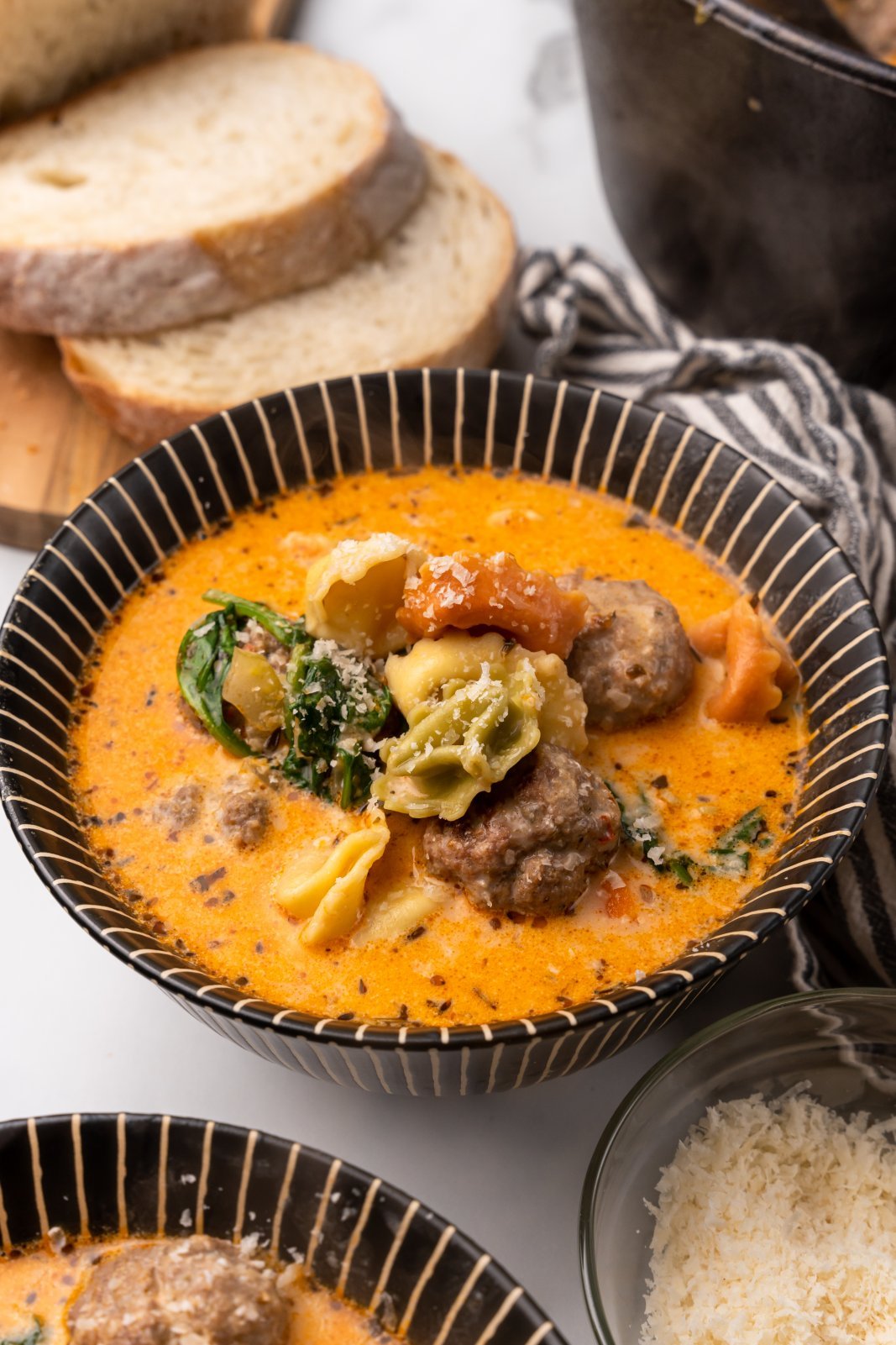 a large bowl of meatball tortellini soup is shown with parmesan sprinkled on top and sliced bread in the background