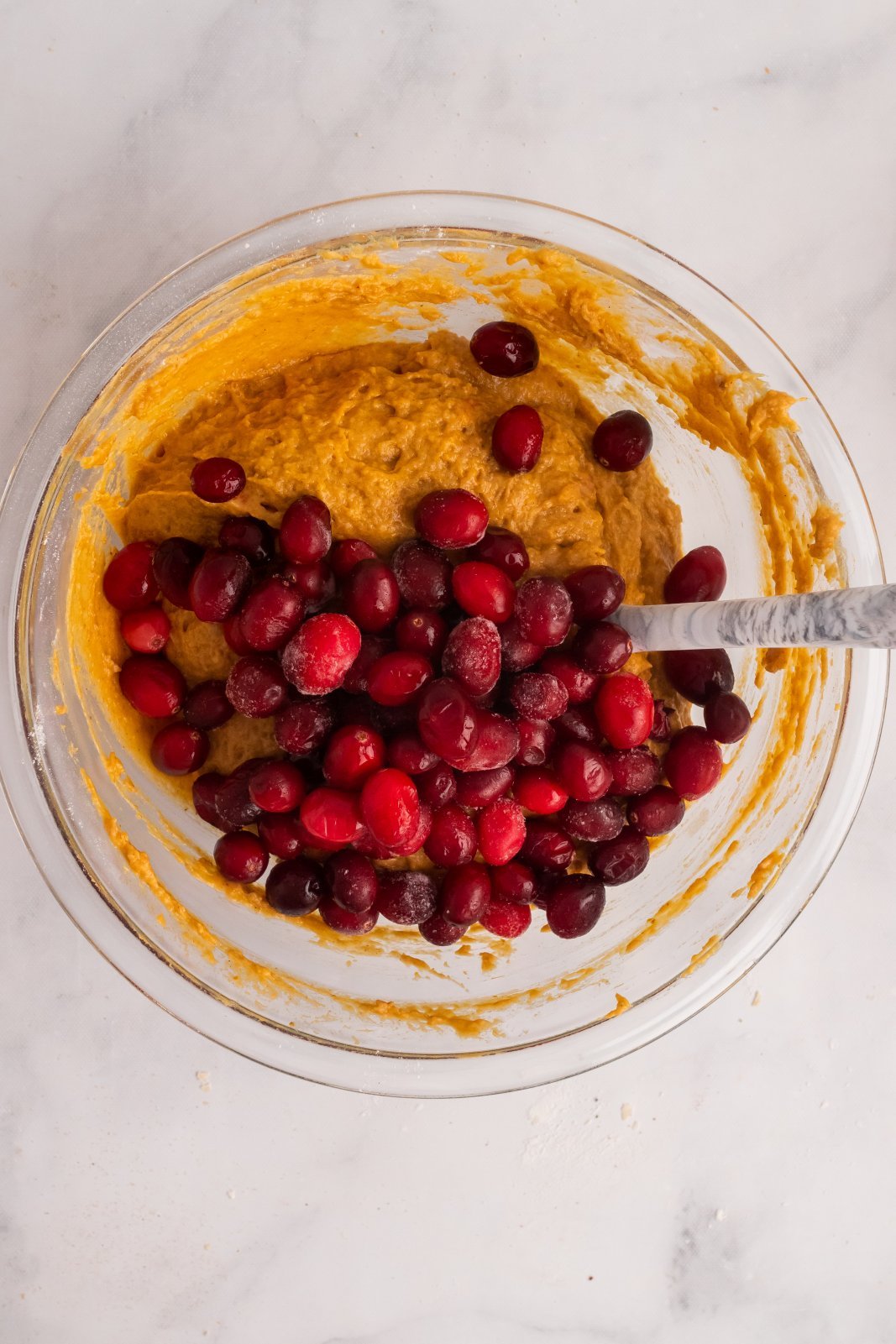cranberries are shown on top of pumpkin muffin batter with a spoon propped in the bowl about to stir
