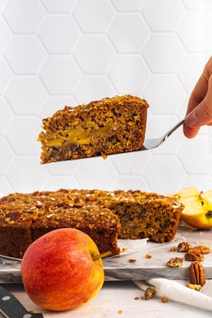 a hand uses a cake server to lift a slice of apple oatmeal cake up