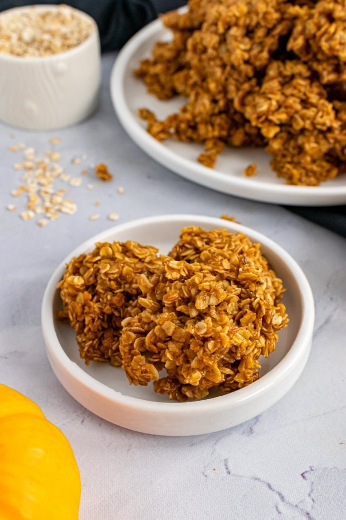 three no bake pumokin spice cookies on a small plate with a pumpkin and a bowl of oats just out of view