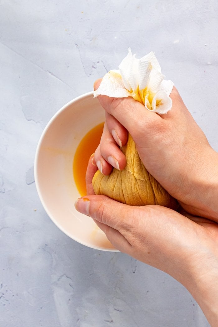 hands squeeze pumpkin puree wrapped in a paper towel; below you can see orange pumpkin juice in a bowl